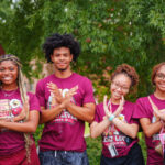 Four young individuals in matching maroon shirts smile and pose with hand gestures in front of a campus signpost surrounded by greenery.