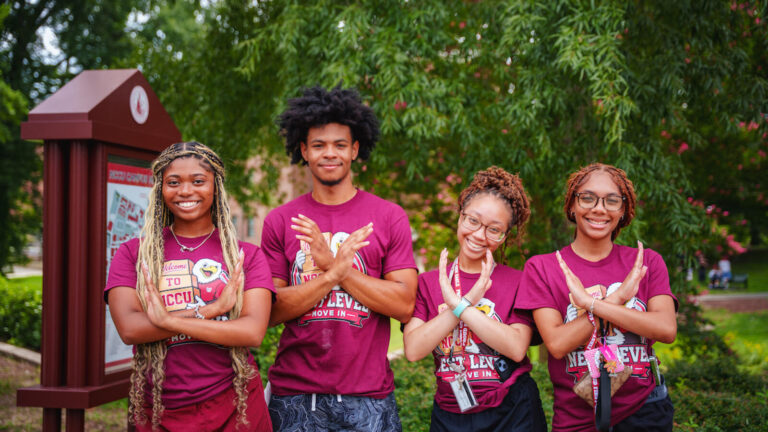 Four young individuals in matching maroon shirts smile and pose with hand gestures in front of a campus signpost surrounded by greenery.