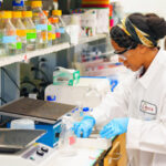 A scientist in a lab coat and gloves works with equipment while another person observes in the background.