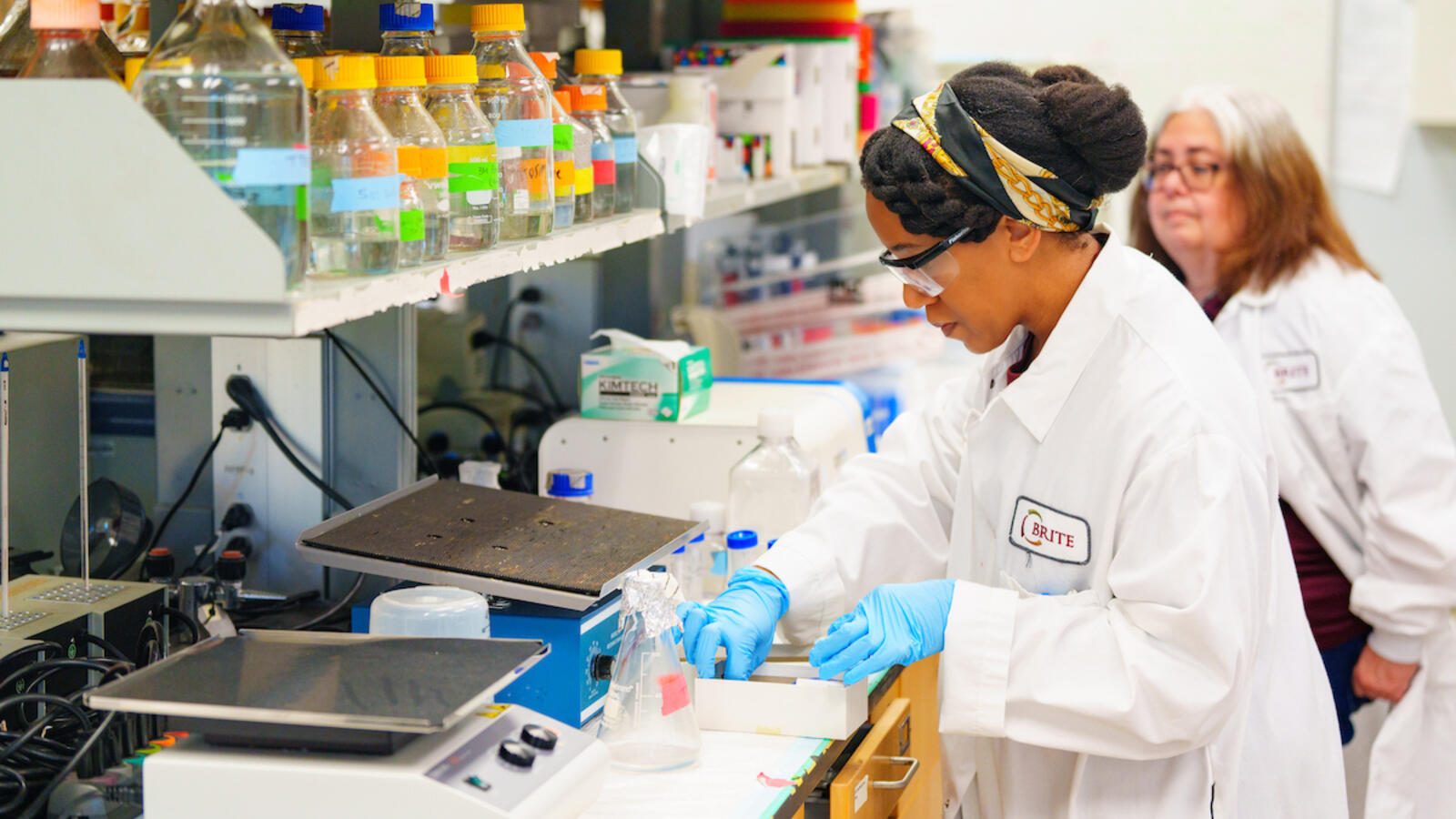 A scientist in a lab coat and gloves works with equipment while another person observes in the background.