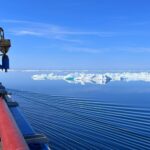 A research vessel sails through calm, reflective Arctic waters dotted with icebergs under a clear blue sky.