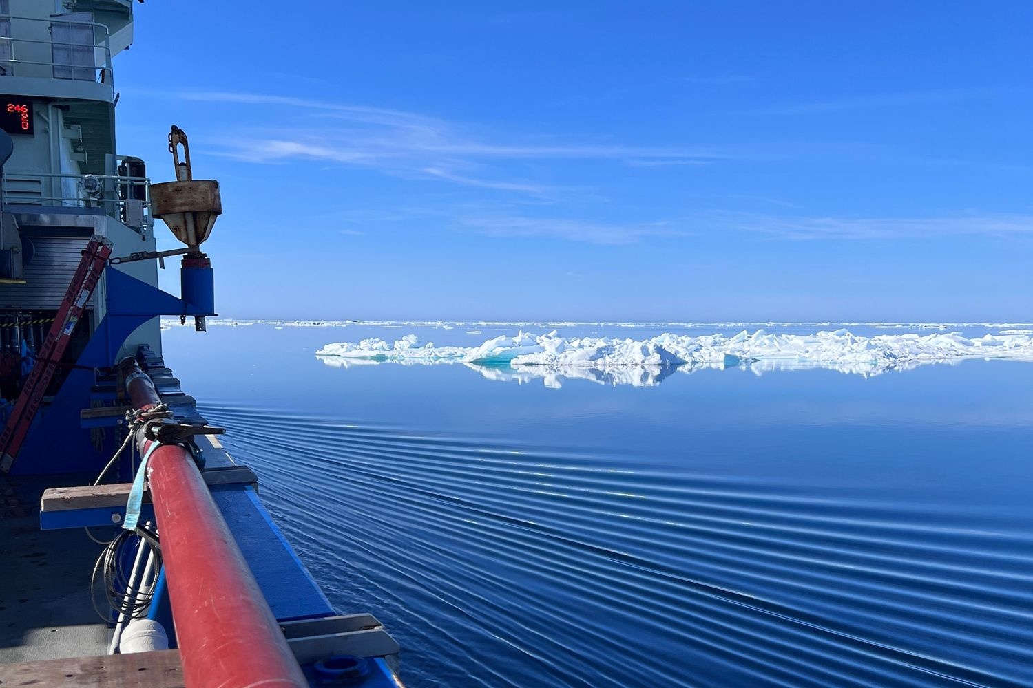 A research vessel sails through calm, reflective Arctic waters dotted with icebergs under a clear blue sky.