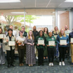 A group of individuals in a room hold certificates and trophies, posing for a photo with smiles and celebratory expressions.
